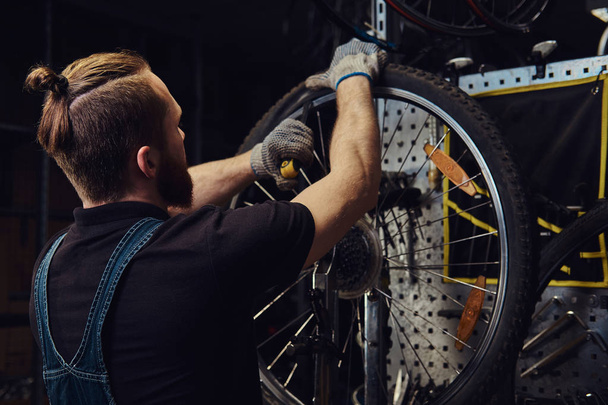 Handsome redhead male in a jeans coverall, working with a bicycle wheel in a repair shop. A worker removes the bicycle tire in a workshop. - Photo, Image