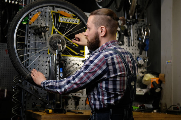 schöner, stylischer Mann in Flanellhemd und Jeans-Overall, der in einer Werkstatt mit einem Fahrradrad arbeitet. - Foto, Bild
