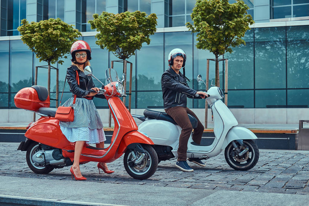 Attractive romantic couple, a handsome man and sexy female, sitting on retro Italian scooters against a skyscraper. - Photo, Image