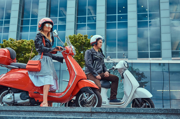 Attractive romantic couple, a handsome man and sexy female, sitting on retro Italian scooters against a skyscraper. - Photo, Image