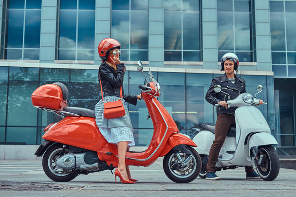 Attractive romantic couple, a handsome man and sexy female sitting on retro Italian scooters against a skyscraper. A man waiting while girl talking by phone. - Photo, image
