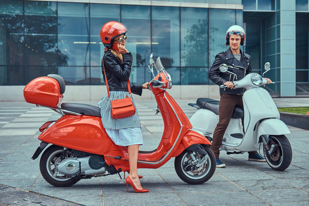 Attractive romantic couple, a handsome man and sexy female sitting on retro Italian scooters against a skyscraper. A man waiting while girl talking by phone. - Photo, Image