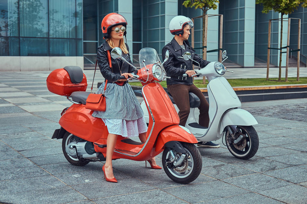 Atractiva pareja romántica, un hombre guapo y sexy hembra, de pie con dos scooters italianos retro contra un rascacielos
. - Foto, Imagen