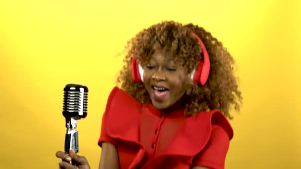 African american girl in red headphones singing in retro microphone. Yellow background - Πλάνα, βίντεο