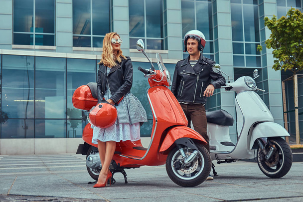 Aantrekkelijke romantisch paar, een knappe man en sexy vrouw, permanent met twee retro Italiaanse scooters tegen een wolkenkrabber. - Foto, afbeelding