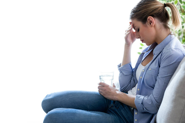 Unhealthy young woman with headache holding glass of water while sitting on sofa at home. - Foto, imagen