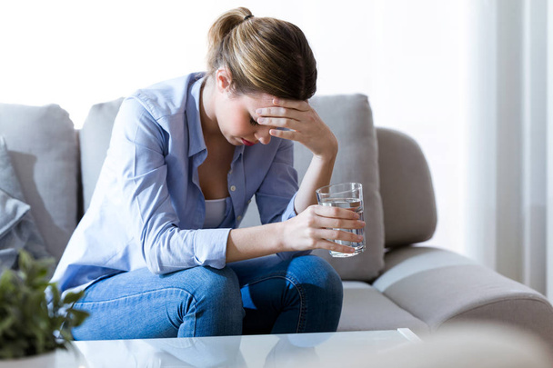 Unhealthy young woman with headache holding glass of water while sitting on sofa at home. - Photo, Image