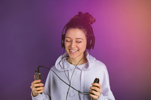 Smiling young beautiful girl with white teeth listening to music on the phone wearing headphones in a sweatshirt on a purple background, toning - Photo, Image