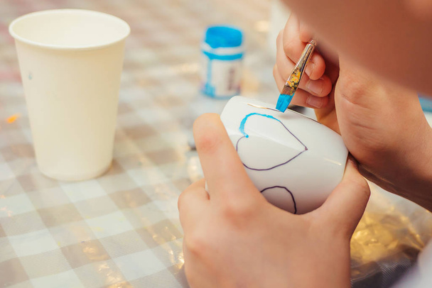A boy draws a heart on the white cup with a blue paint  - Photo, Image
