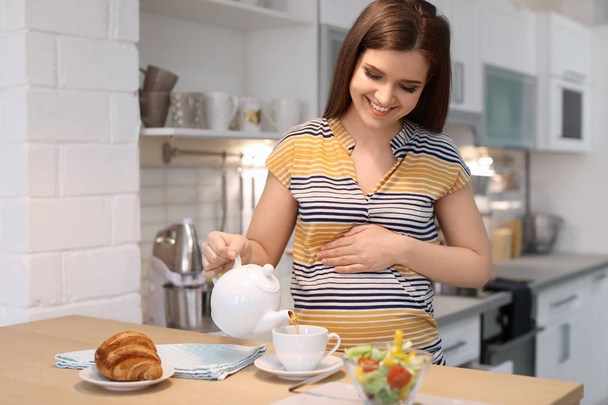 Young pregnant woman pouring tea into cup at table in kitchen - Photo, image