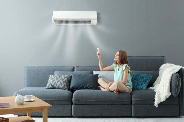 Young woman switching on air conditioner while sitting on sofa at home - Photo, Image