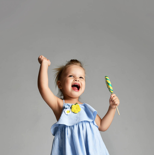 Young pretty toddler girl kid with big  sweet lollypop candy in blue dress on grey - Фото, зображення