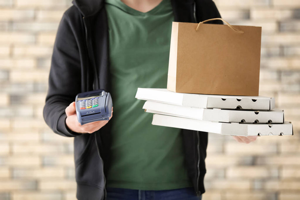 Молодой человек с картонными коробками для пиццы, бумажным пакетом и банковским терминалом на фоне кирпичной стены. Доставка продуктов питания
 - Фото, изображение