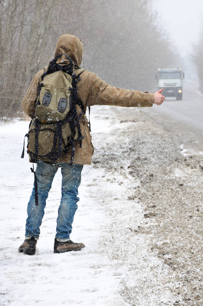 ein Mann mit einem großen Rucksack, der Daumen nach oben zeigt, während er im winterlichen Schneesturm eine Asphaltstraße entlang trampt - Foto, Bild
