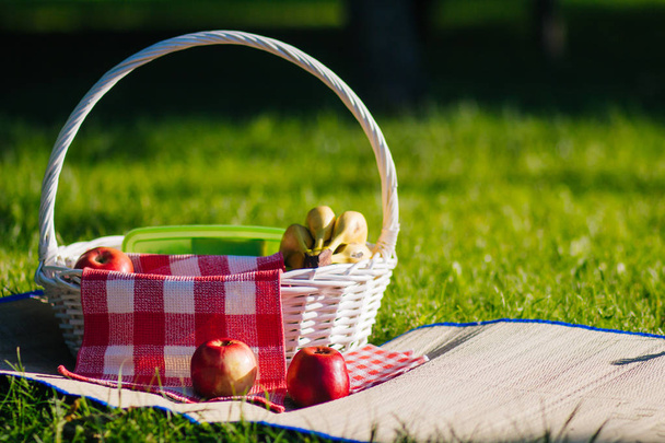 Білий фруктовий кошик стоїть на соломі. Рушник у червоній клітці. Пікнік на газоні. Букет жовтих бананів і червоних яблук
 - Фото, зображення