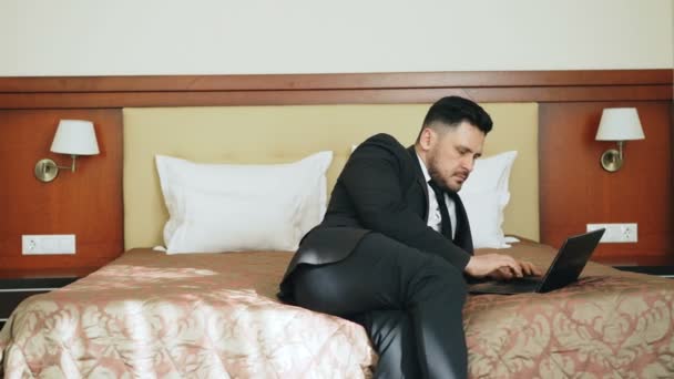 Pan shot of concentrated businessman in suit working on laptop while sitting on bed in hotel room. Business, travel and people concept - Imágenes, Vídeo