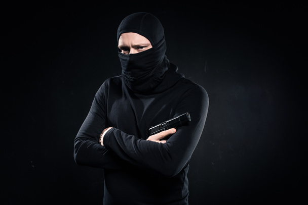 Zloděj v černou kuklu drží zbraň v založenýma rukama na černém pozadí - Fotografie, Obrázek