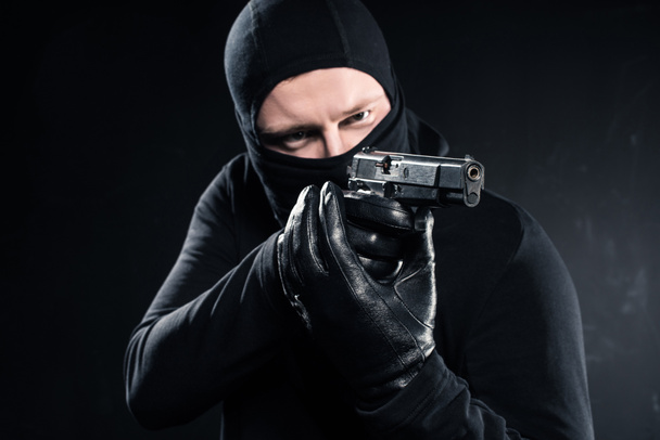 Hombre ladrón en pasamontañas apuntando con arma en negro
 - Foto, Imagen
