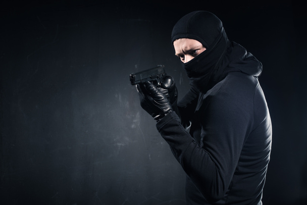 Homme criminel en cagoule et gants visant avec arme à feu sur noir
 - Photo, image