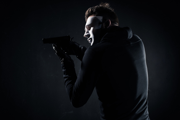 Ladrón en máscara y guantes apuntando con pistola en negro
 - Foto, Imagen