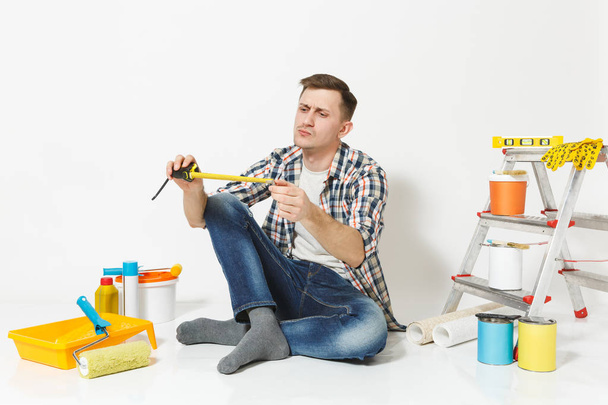 Man in casual clothes sitting on floor with measure tape, instruments for renovation apartment room isolated on white background. Wallpaper, gluing accessories, painting tools. Repair home concept. - Photo, Image