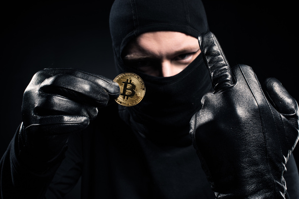 Man in black balaclava holding golden bitcoin and showing middle finger - Fotó, kép