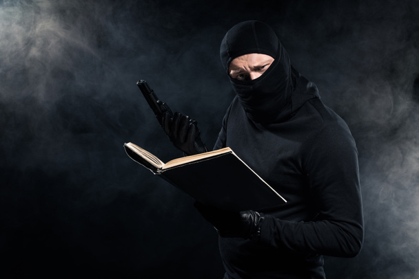 Man in zwarte balaclava holding pistool en leesboek - Foto, afbeelding