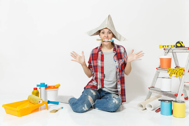 Woman in newspaper hat sitting on floor with brush in mouth, instruments for renovation apartment isolated on white background. Wallpaper, accessories for gluing, painting tools. Repair home concept. - Photo, Image