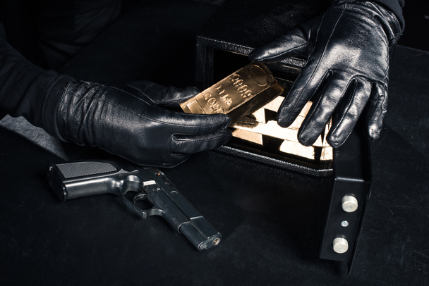 Крупный план грабителя с пистолетом, забирающего золотые слитки из сейфа
 - Фото, изображение