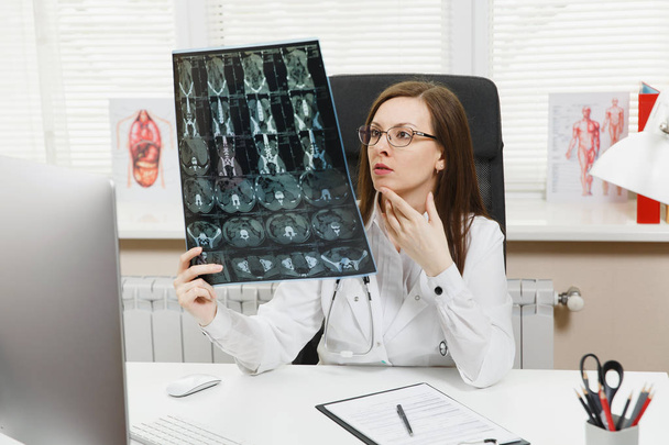 Vrouwelijke arts zit aan bureau met computer, film x-ray de hersenen door radiografisch beeld ct scan mri in lichte kantoor in ziekenhuis. Vrouw in medische jurk in raadpleging van de kamer. Gezondheidszorg, geneeskunde concept. - Foto, afbeelding