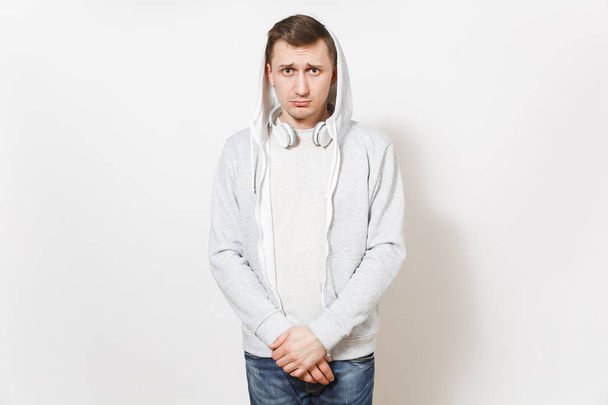 Young handsome sad upset man student in t-shirt, blue jeans and light sweatshirt with hood with headphones with guilty face folds his arms isolated on white background. Concept of emotions - Photo, Image
