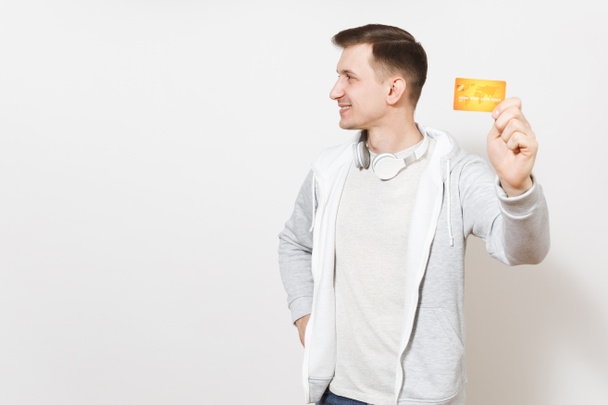 Jeune bel homme souriant étudiant en t-shirt, sweat-shirt léger avec casque autour du cou montre la carte de crédit orange avec de l'argent à la caméra, regarde loin isolé sur fond blanc. Concept de succès
 - Photo, image