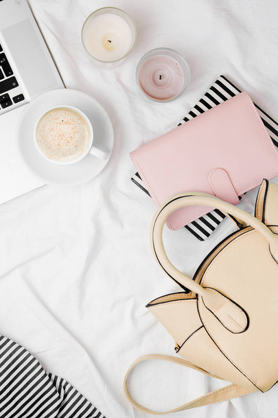 Fashion blogger workspace with laptop, beige handbag, coffee, candles and notebooks in bed. - Photo, image