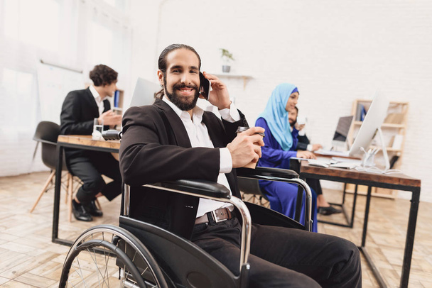 Disabled arab man in suit in wheelchair talking on smartphone and drinking coffee - Fotografie, Obrázek