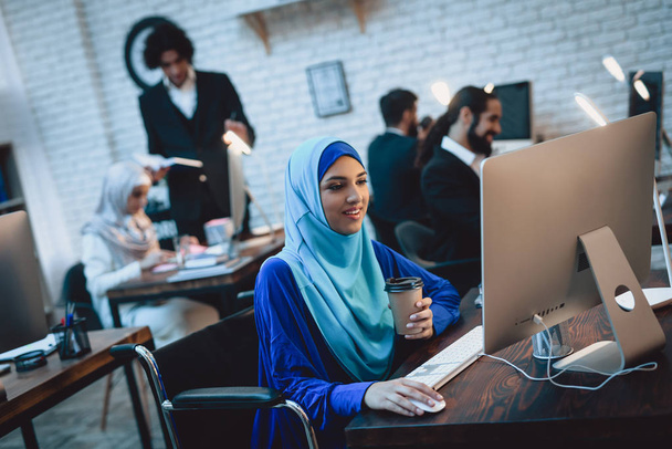 Disabled arab woman in hijab in wheelchair working on desktop computer and drinking coffee - Фото, изображение