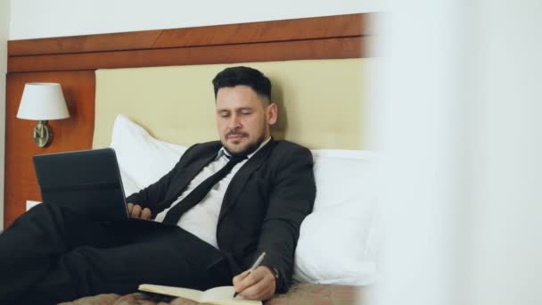 Confident businessman lying on bed using laptop computer and writing notes at notepad while working in hotel room. Travel, business and people concept - Video