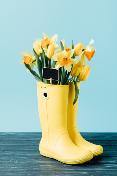 close up view of yellow tulips and narcissus flowers with empty chalkboard in rubber boots on blue - Foto, Imagem