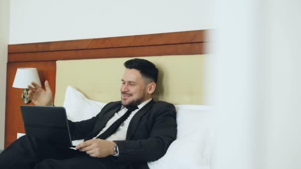 Happy businessman talking with family via online video chat using laptop computer lying on bed in hotel room. Travel, business and people concept - Materiał filmowy, wideo