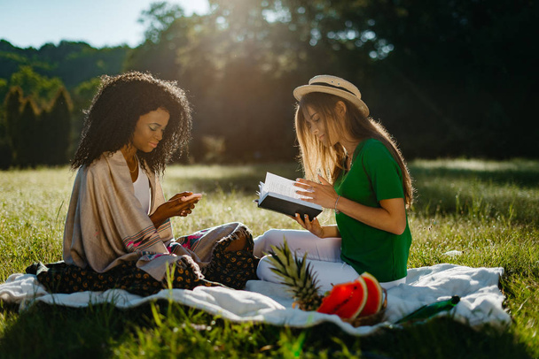 Duas amigas multi-étnicas durante o piquenique no prado ensolarado. A menina africana atraente está conversando e navegando através do telefone celular, enquanto sua linda amiga loira está lendo o livro
. - Foto, Imagem