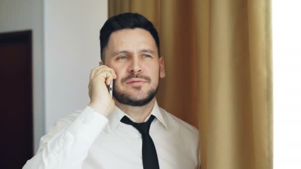 Взрослый улыбающийся бизнесмен в белой рубашке и галстуке разговаривает по мобильному телефону, стоя у окна в номере отеля
 - Кадры, видео