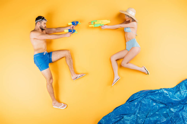 junges Paar in Badebekleidung spielt mit Wasserpistolen auf gelb - Foto, Bild
