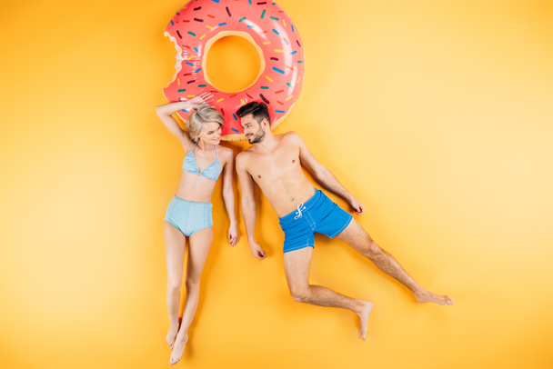 feliz joven pareja en traje de baño sonriendo el uno al otro mientras está acostado en el anillo de natación en amarillo
 - Foto, imagen