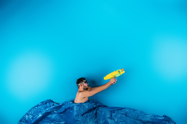 young man in swimming mask shooting with water gun from imagine waves on blue - Foto, afbeelding