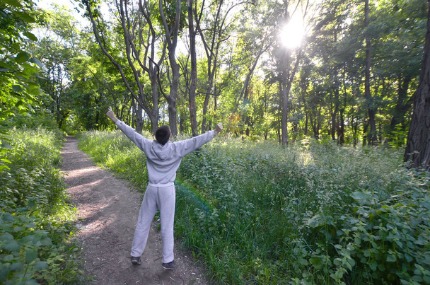 A young guy in a gray sports suit rejoices in the rising of the sun among the trees in the forest. Recreation during a sports run in the open air forest. The delight of a beautiful dawn - Photo, Image