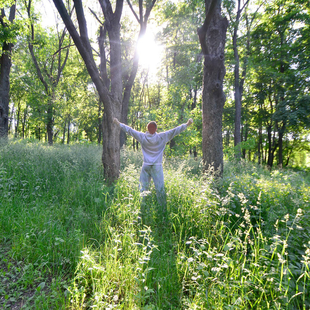 Молодой парень в сером спортивном костюме радуется восходу солнца среди деревьев в лесу. Отдых во время спортивных пробежек в лесу под открытым небом. Восхищение прекрасным рассветом
 - Фото, изображение