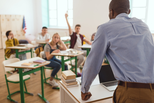 Rear view of african american teacher and schoolboy with arm up in classroom  - Foto, imagen