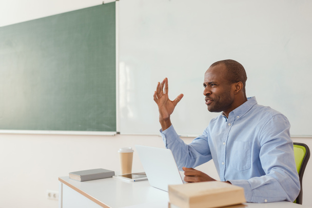Frusztrált afro-amerikai tanár intett, és ül íróasztal, laptop  - Fotó, kép