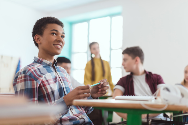 Vista de ángulo bajo del estudiante afroamericano de secundaria sosteniendo lápiz en la mano y compañeros de clase sentados detrás
 - Foto, Imagen