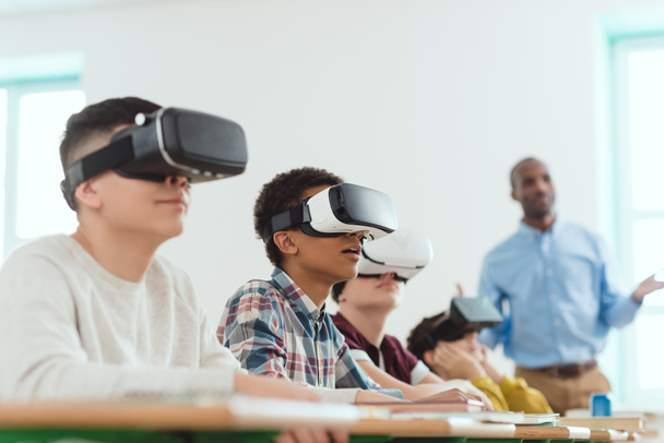 scolari multiculturali utilizzando cuffie realtà virtuale e insegnante afro-americano in piedi dietro
 - Foto, immagini