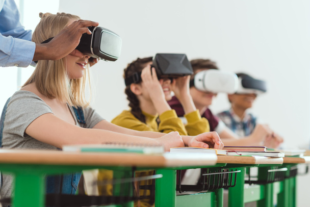 Imagen recortada del profesor afroamericano ajustando los auriculares de realidad virtual a la colegiala adolescente con compañeros sentados detrás
 - Foto, Imagen